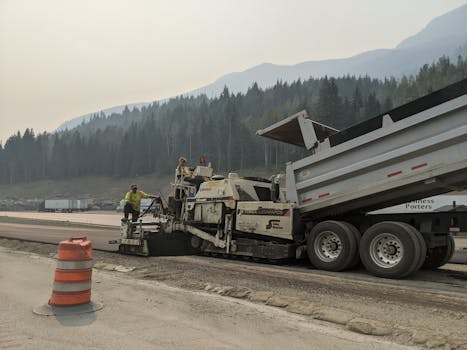 Road workers paving a highway amidst mountainous forest scenery.
